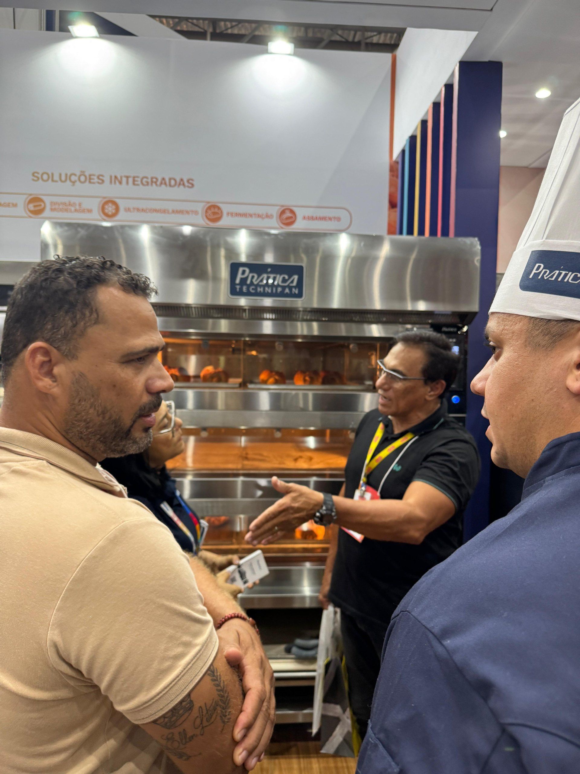 Pessoas conversando perto de um forno industrial em uma feira de negócios; o forno está assando alimentos.