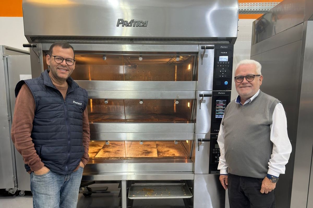 Dois homens em pé perto de um forno comercial; um usa óculos e um colete, o outro, uma jaqueta.