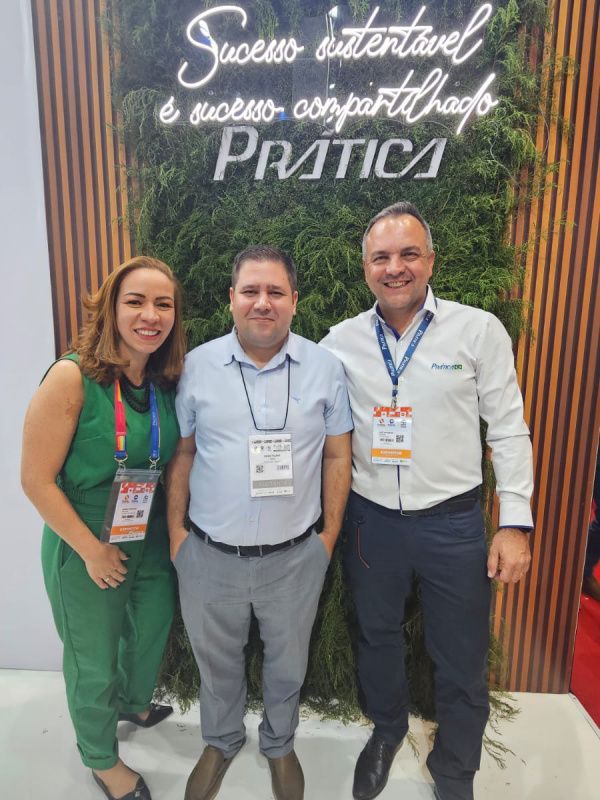 Três pessoas estão posando para uma foto em frente a uma placa que diz pratica