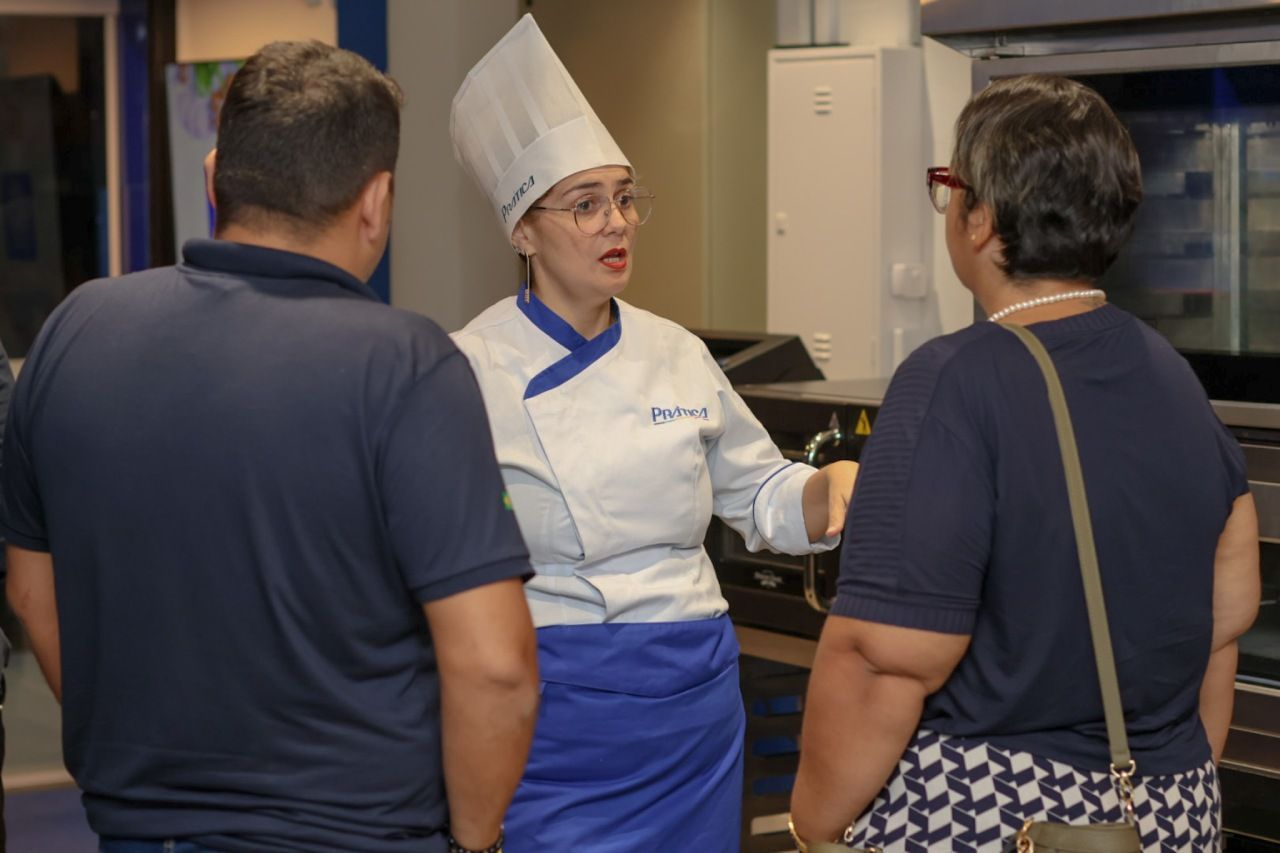 Um chefe de cozinha uniformizado conversando com duas pessoas em uma cozinha industrial.