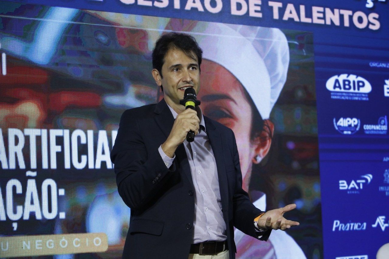 Um homem de terno segura um microfone em frente a uma tela grande.