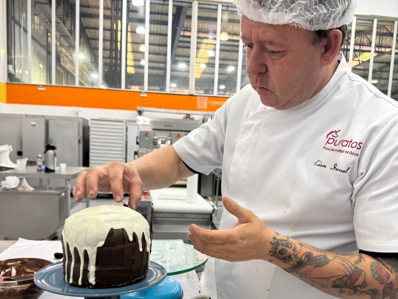 Em uma padaria, um padeiro de jaleco branco e touca examina um bolo de chocolate com gotas de glacê branco.