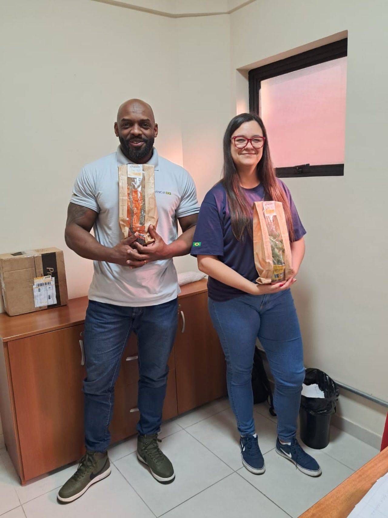 Um homem e uma mulher estão de pé, um ao lado do outro, em uma sala, segurando sacos de comida.