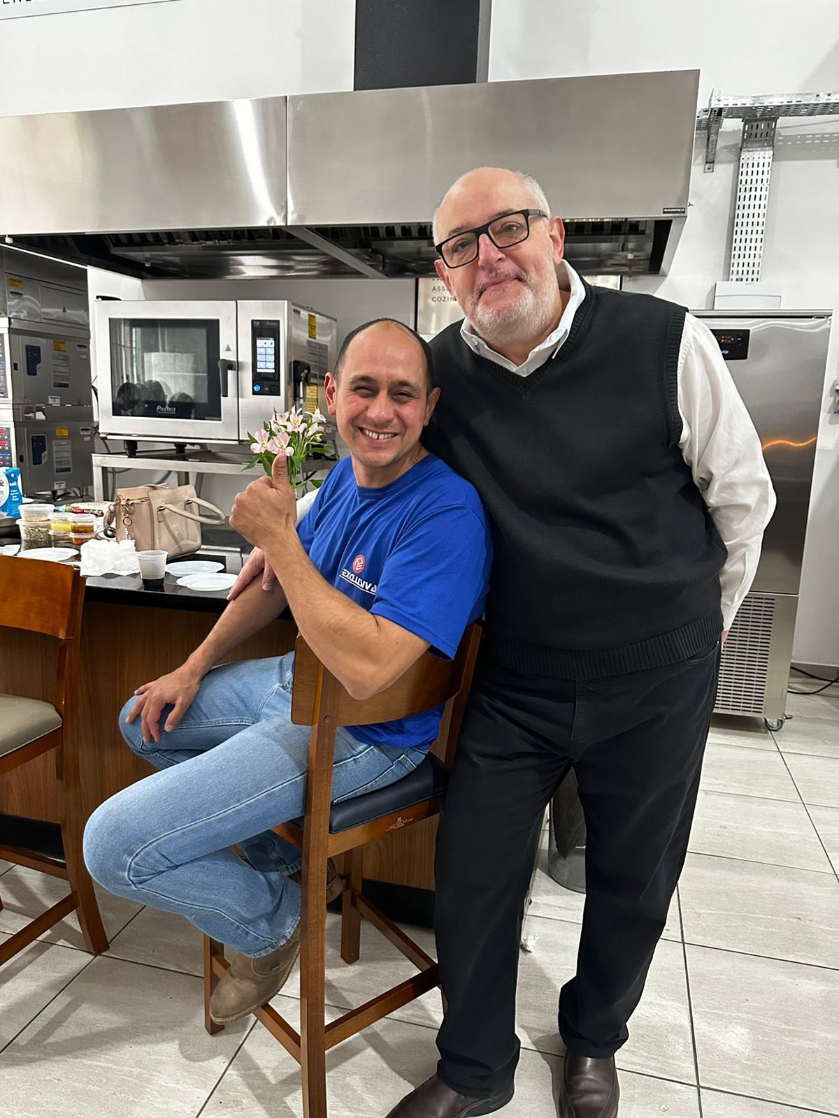 Dois homens estão posando para uma foto em uma cozinha. um está sentado em uma cadeira e o outro está de pé.