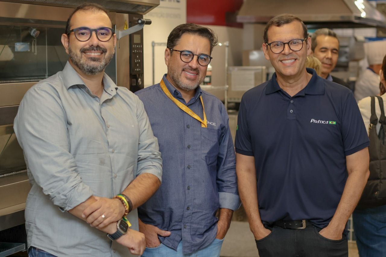 Três homens sorridentes, de pé próximos um do outro em uma cozinha comercial, usando óculos e roupas casuais.