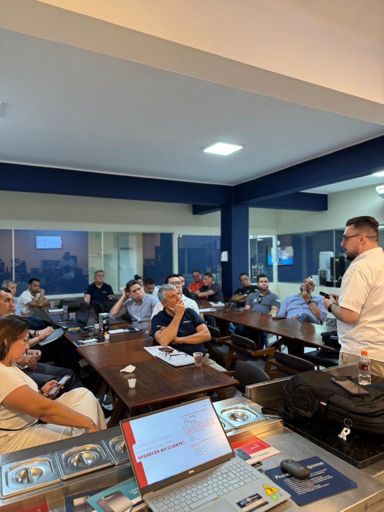 Homem fazendo uma apresentação para um grupo sentado à mesa em uma sala. Laptop, papéis e refrescos estão sobre uma mesa.