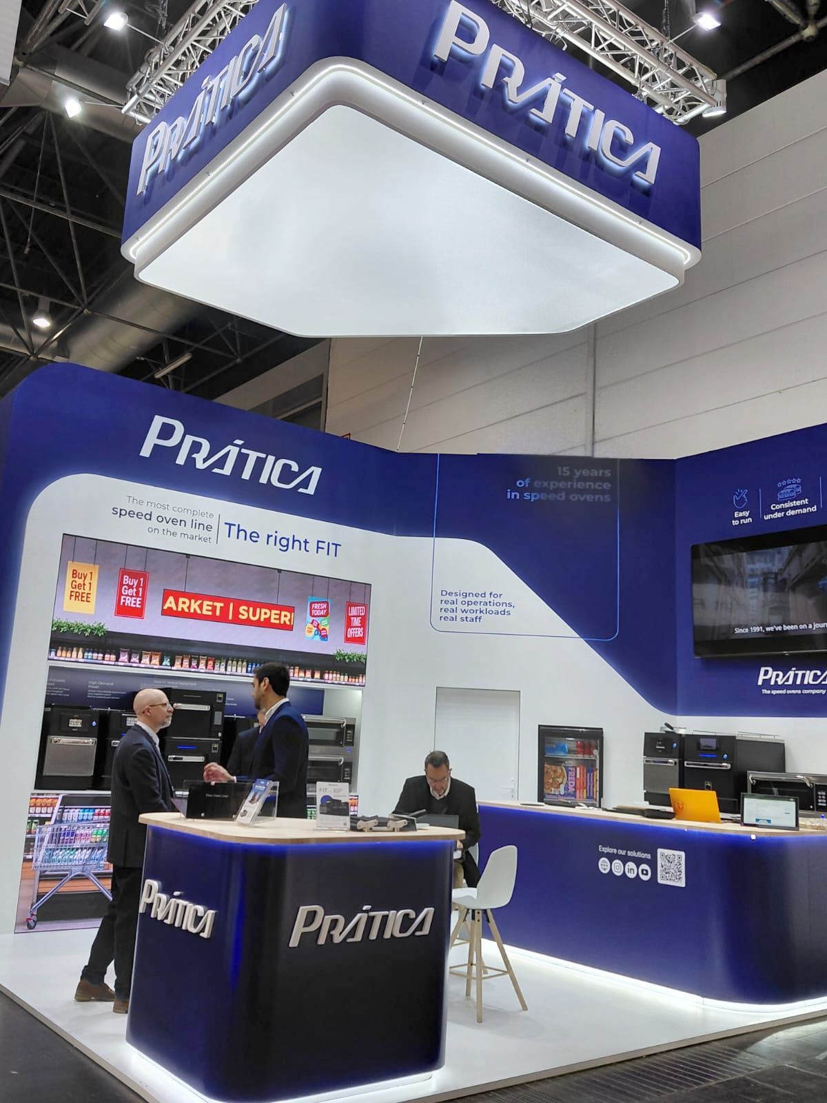Estande da Prática em feira comercial, azul e branco, com funcionários interagindo e exposição de produtos.