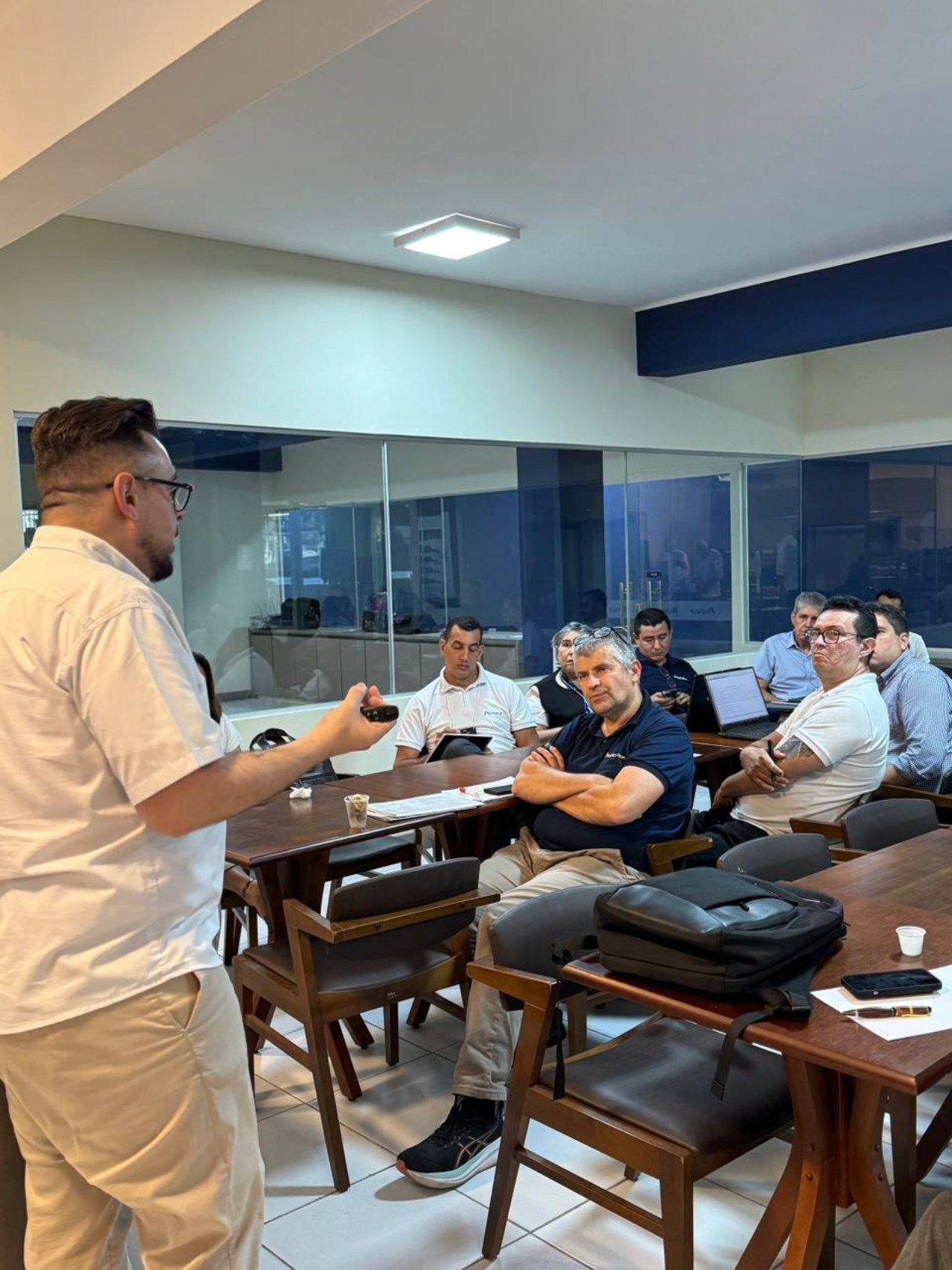 Homem apresentando em uma sala de reuniões, gesticulando em direção a um grupo de pessoas sentadas; período diurno.