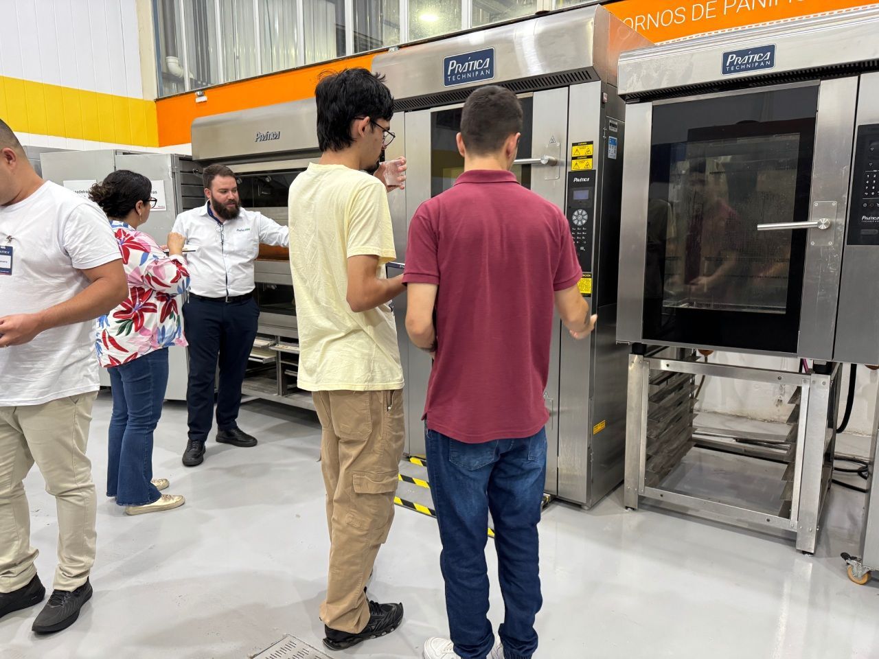 Pessoas observando fornos industriais em uma loja. Um homem com uma camisa bordô examina um deles.