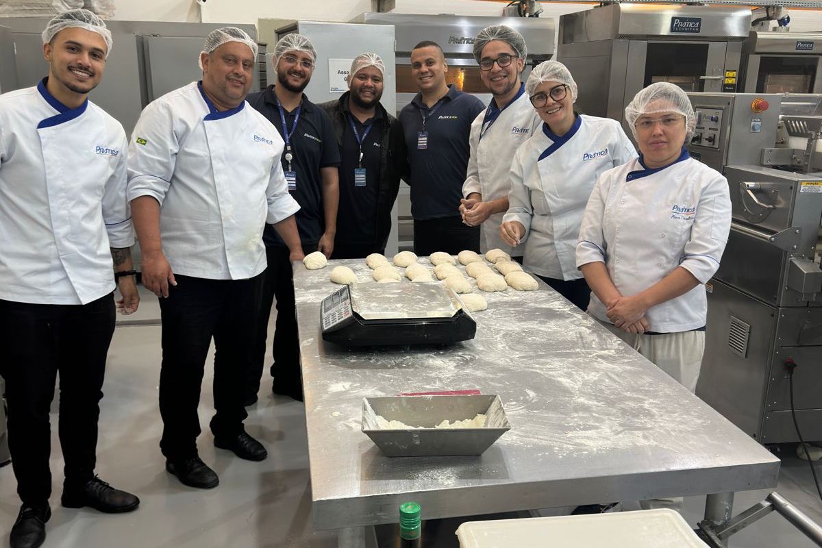 Grupo de chefs em uma cozinha preparando massa, sorrindo. Balcões, fornos e equipamentos de aço inoxidável são visíveis.