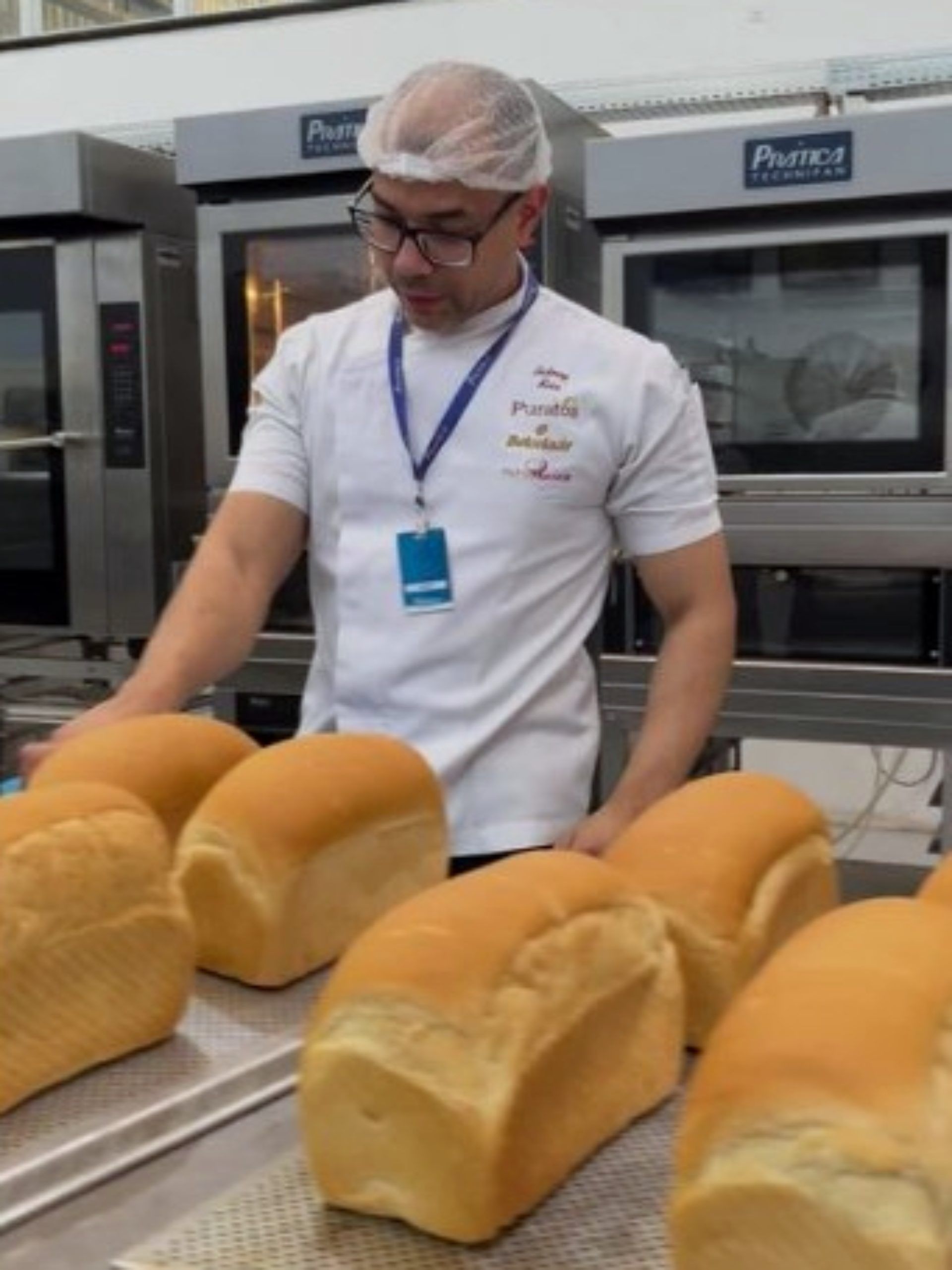 Um homem de uniforme branco está parado em frente a uma fileira de pães