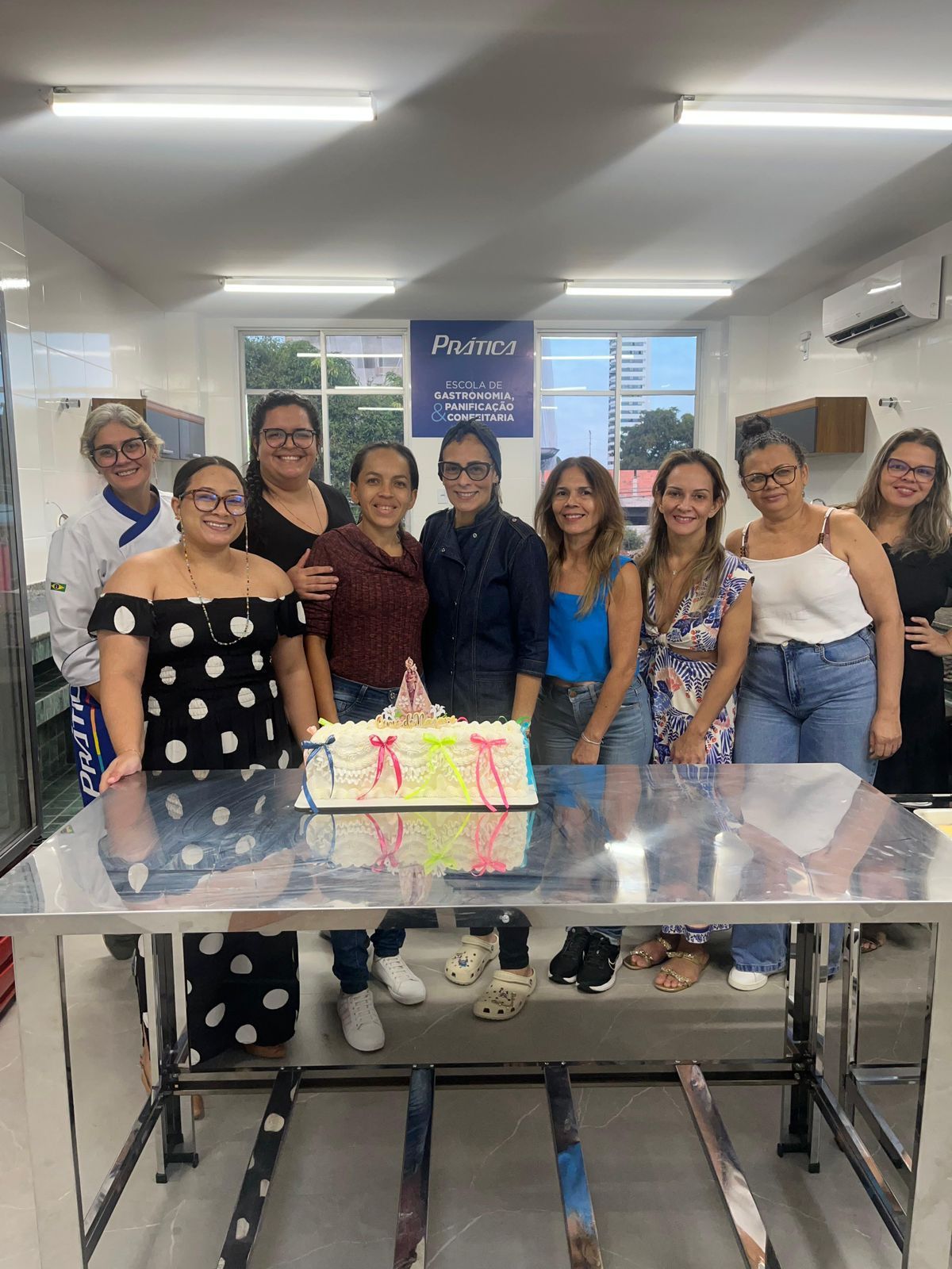 Grupo de mulheres sorrindo ao redor de um bolo sobre uma mesa em um escritório bem iluminado.
