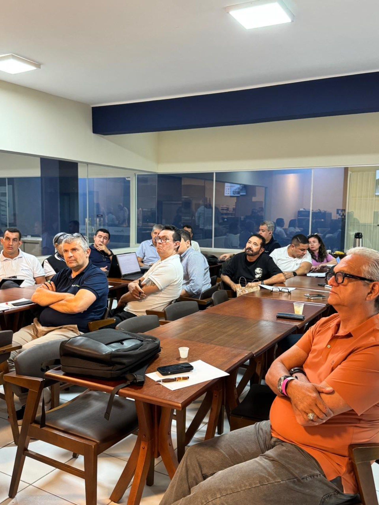 Grupo de pessoas em volta de uma mesa comprida, possivelmente em uma reunião ou apresentação. Algumas olhando para a câmera.