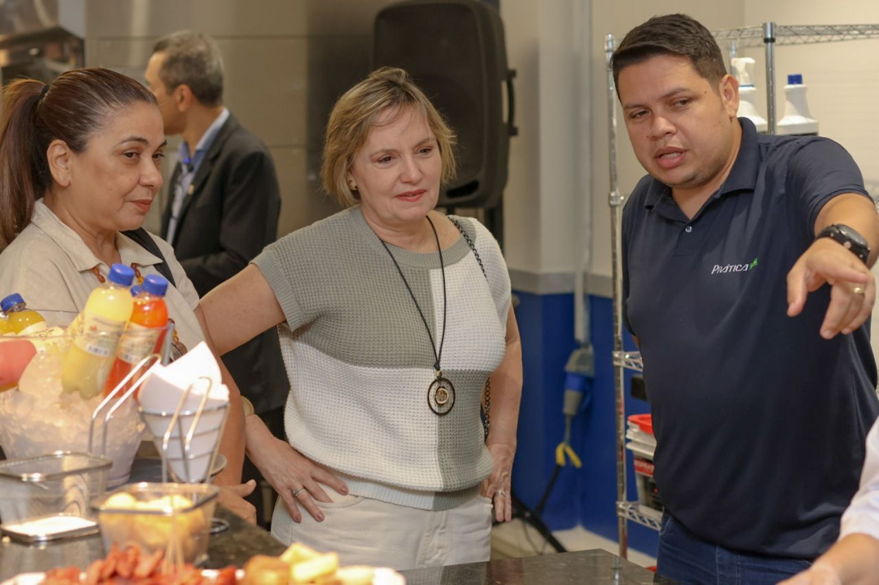 Três pessoas em uma cozinha; um homem aponta, duas mulheres escutam, cercadas por comida e utensílios.