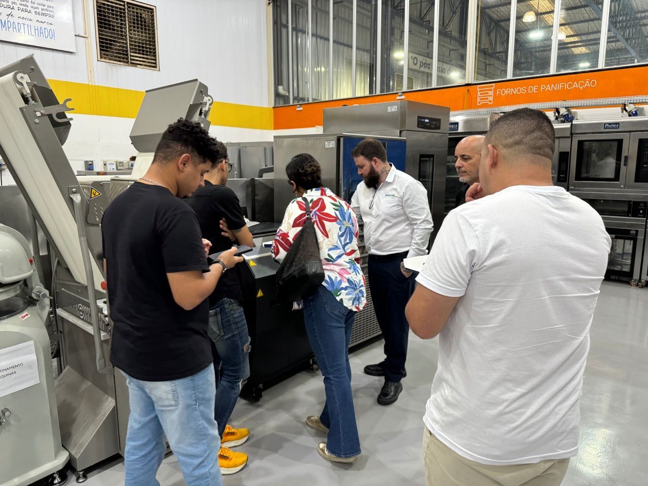 Grupo de pessoas observando máquinas em um ambiente fabril. Uma pessoa de camisa branca parece estar explicando o funcionamento da máquina.