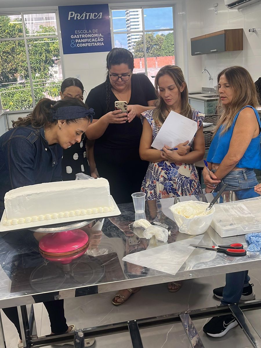 Grupo de mulheres em uma aula de confeitaria, olhando para um bolo coberto, em ambiente iluminado.