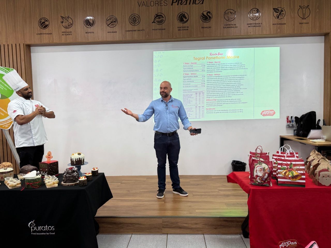 Homem apresentando um chef, uma tela de projeção com texto e bolos sobre as mesas.