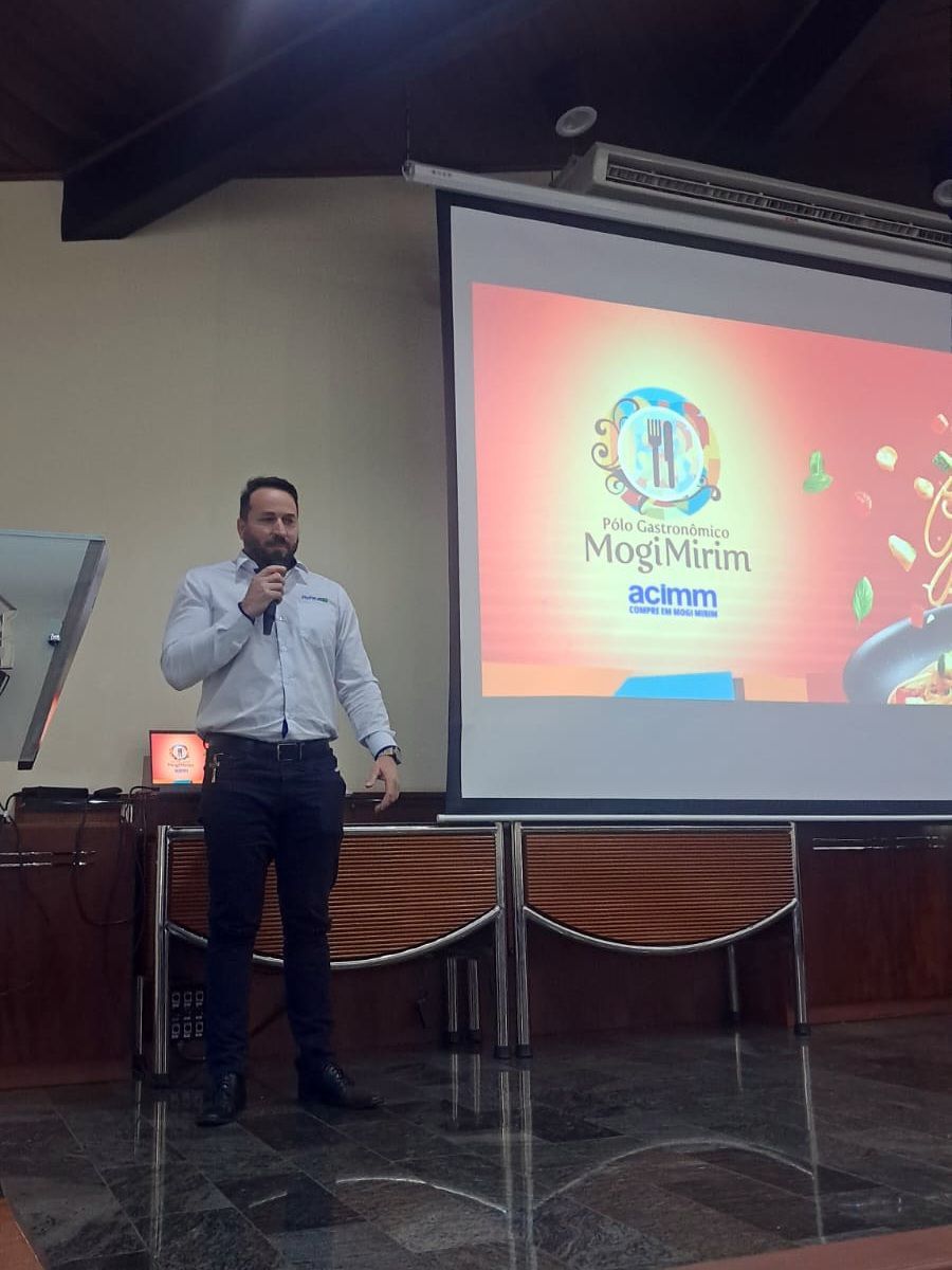Homem fazendo uma apresentação em um salão, com uma tela exibindo um logotipo.