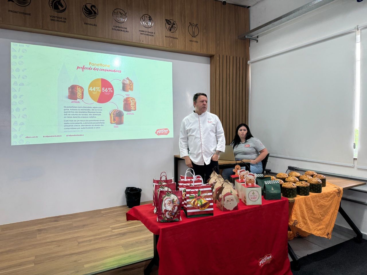 Um homem apresenta um prato perto de uma tela de projeção que exibe diagramas de alimentos. Uma mulher senta-se ao lado dele, e presentes e produtos assados ​​estão dispostos sobre as mesas.
