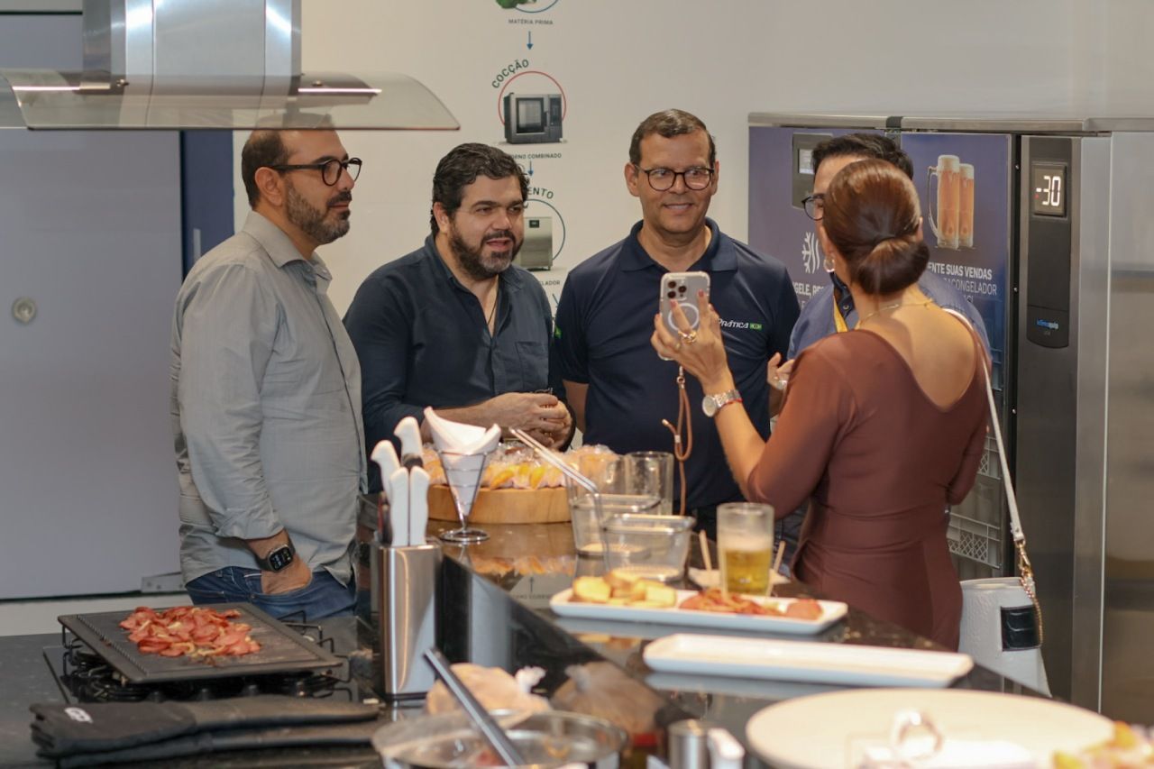 Pessoas tirando uma foto em uma cozinha; uma mulher fotografa três homens reunidos em volta de um bar, e há comida à mostra.