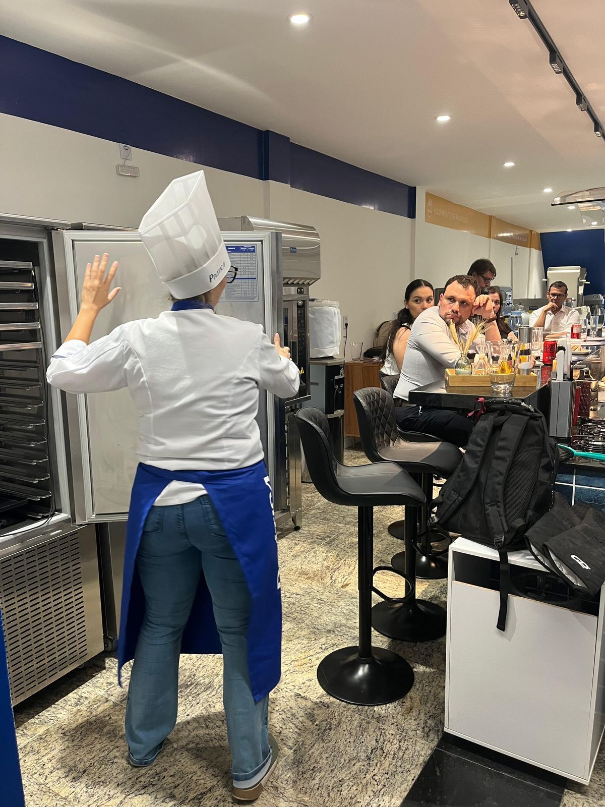 Chef de uniforme branco e avental azul gesticula em direção a uma geladeira e clientes sentados em um bar.