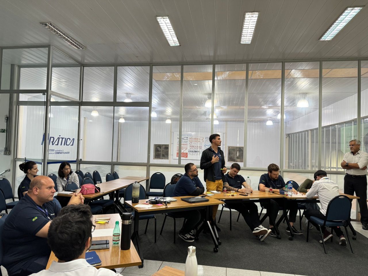 Um grupo de pessoas está reunido em torno de uma mesa em um escritório com paredes de vidro para uma reunião ou workshop.