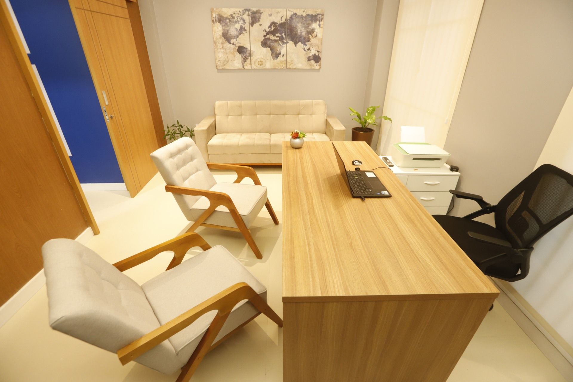 Escritório com mesa, cadeiras, sofá, mapa-múndi e impressora; cores neutras e luz natural.