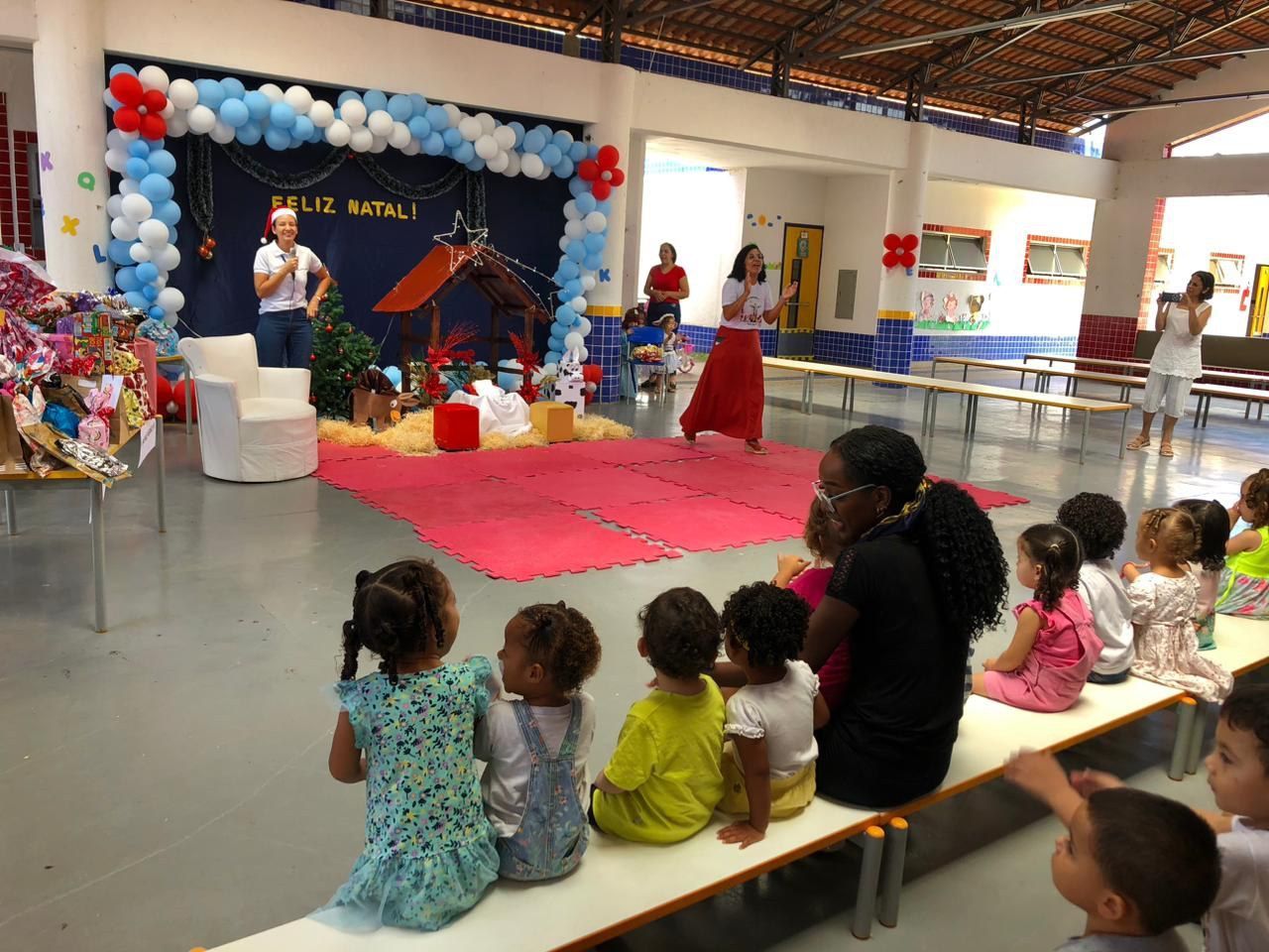 Um grupo de crianças está sentado em bancos em frente a um palco decorado com balões.