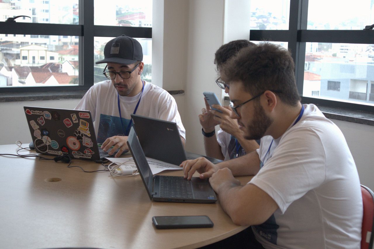 Três homens estão sentados à mesa com laptops e celulares.