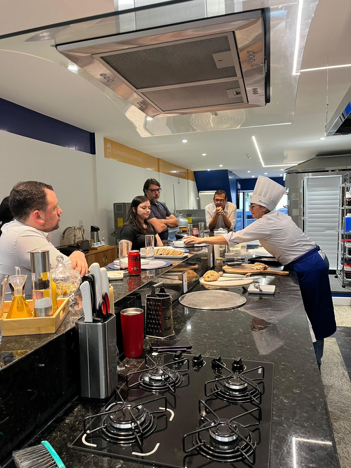 Chef apresentando comida ao grupo em uma cozinha bem iluminada com decoração em azul e branco.