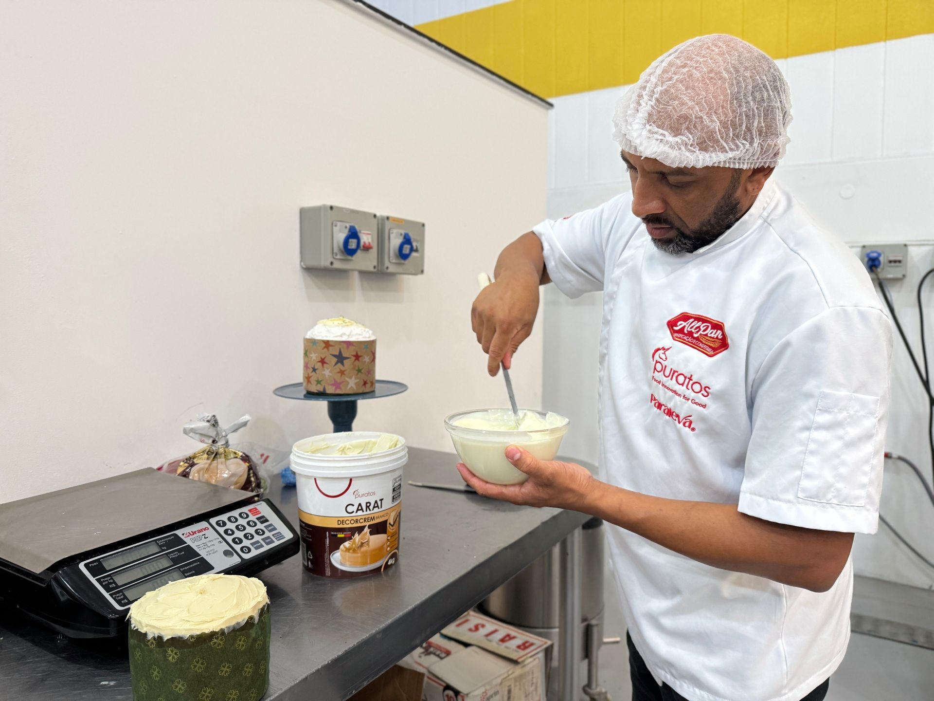 Padeiro mexendo glacê ao lado de bolos e ingredientes já preparados em uma cozinha industrial.