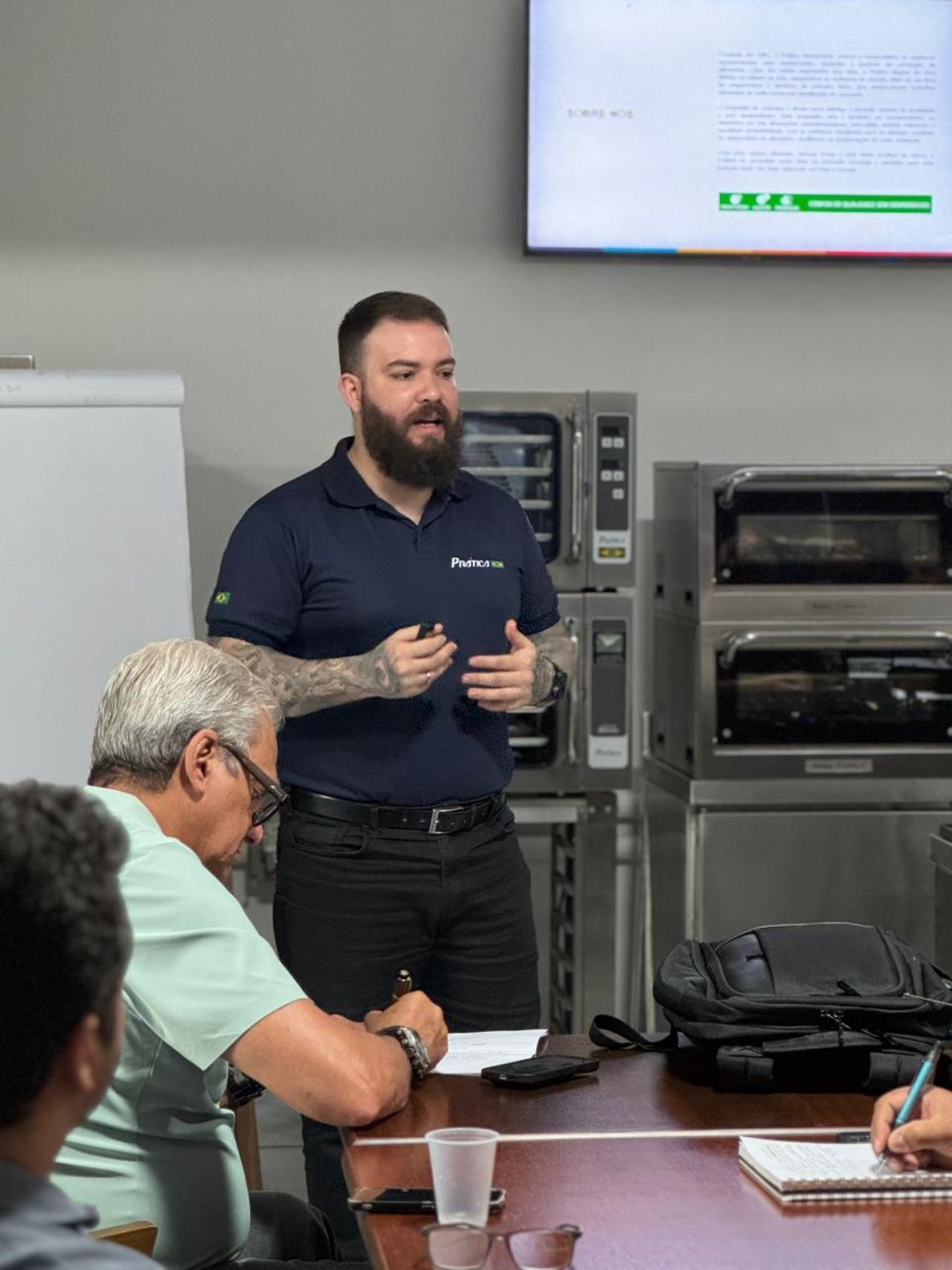 Homem barbudo faz apresentação para um grupo. Apresentação em frente a fornos.