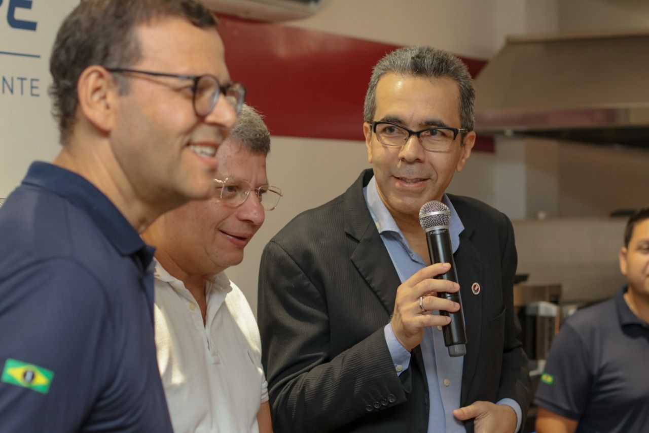 Homem de óculos segurando um microfone, falando para um grupo de homens em um ambiente fechado.