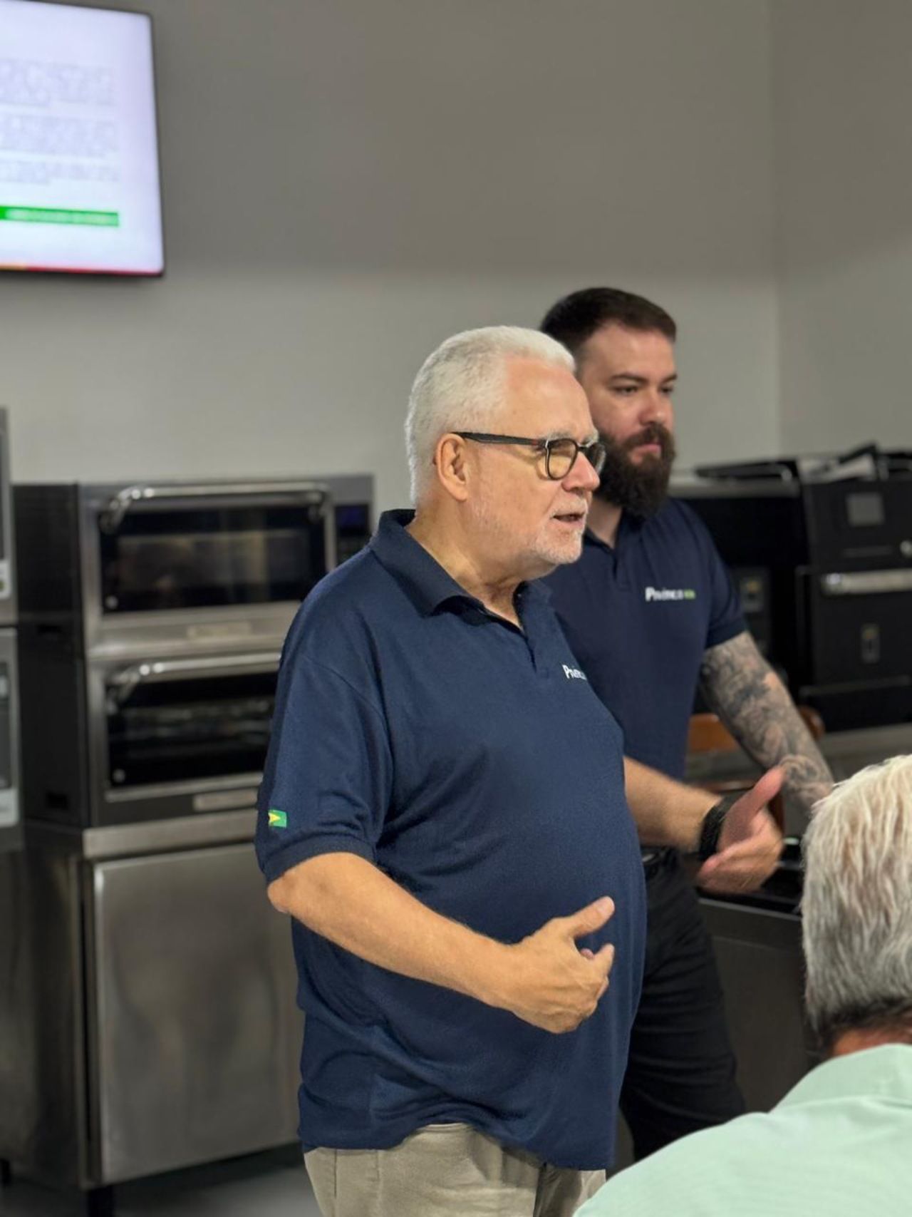 Um homem de camisa azul fala, enquanto outro homem barbudo está atrás dele, perto de fornos industriais.