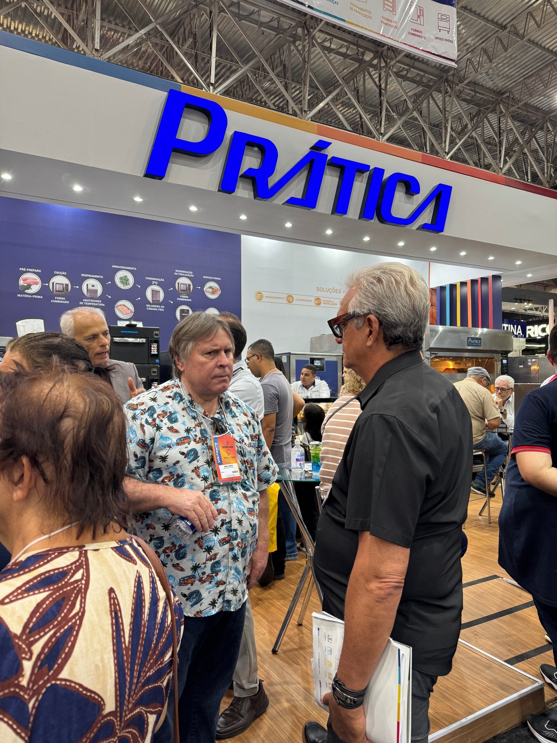 Pessoas conversando no estande da Prática em uma feira comercial. A placa é azul e branca.