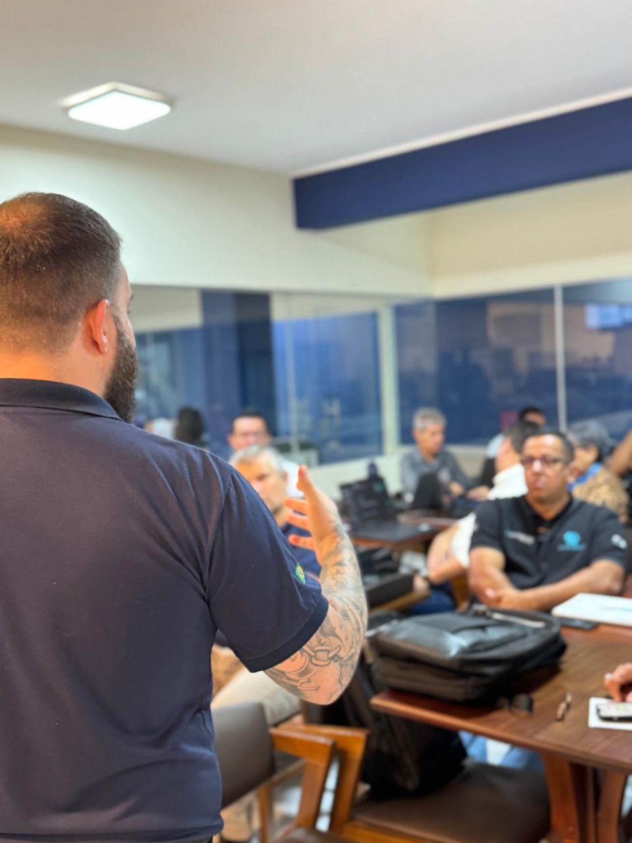 Homem tatuado fazendo uma apresentação em uma sala com um grupo de pessoas sentadas à mesa, fundo desfocado.
