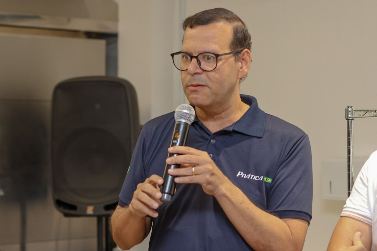 Homem de óculos falando ao microfone, vestindo uma camisa polo azul escura.