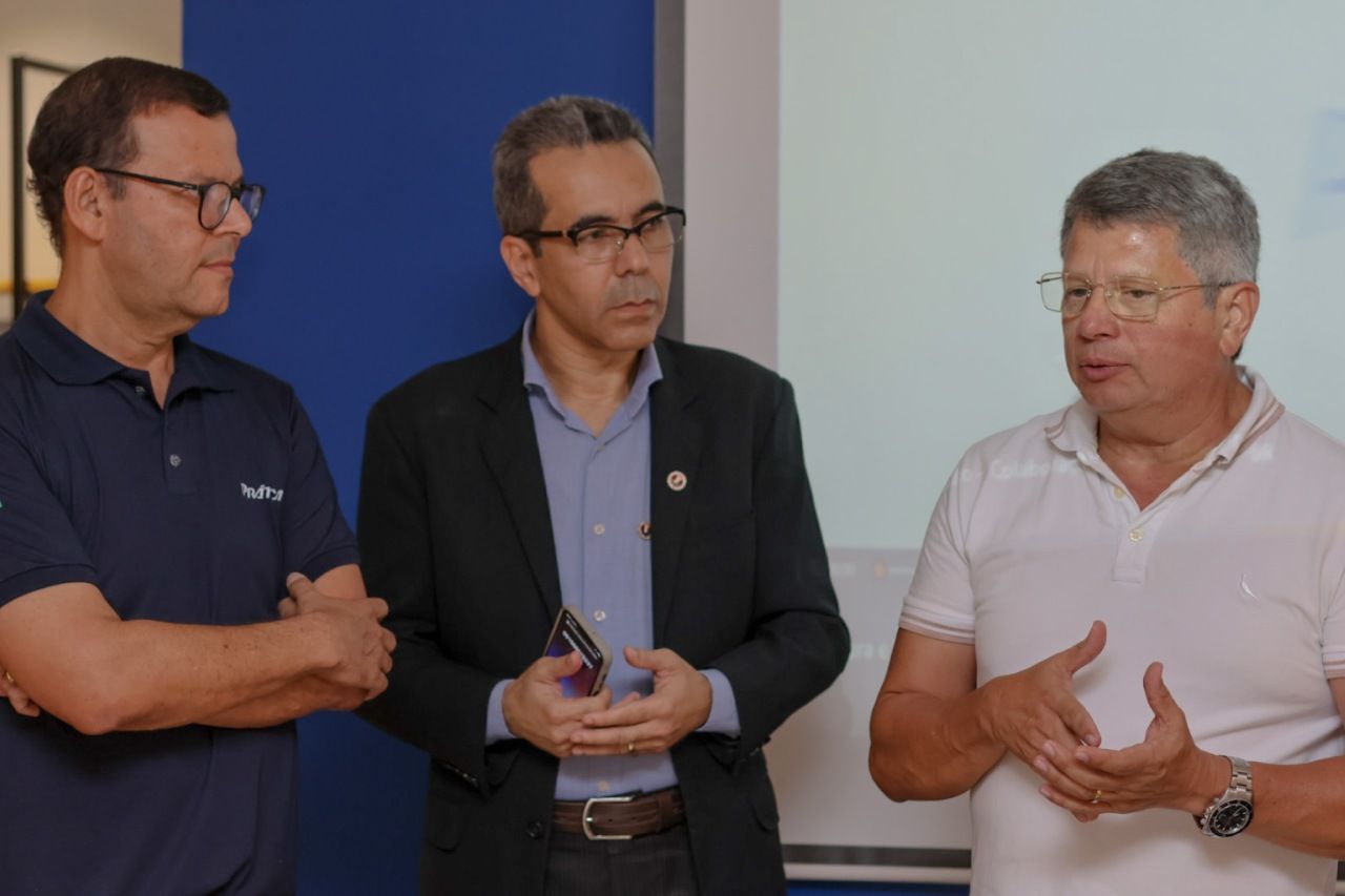 Três homens em uma sala; um falando e gesticulando. Os outros ouvem. Fundo azul e branco.
