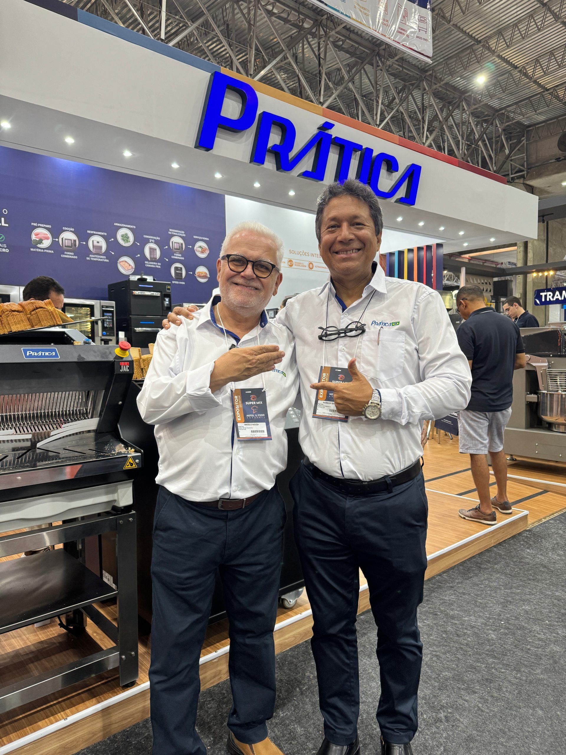 Dois homens sorridentes posando em frente a um estande da “Prática” em uma feira comercial.