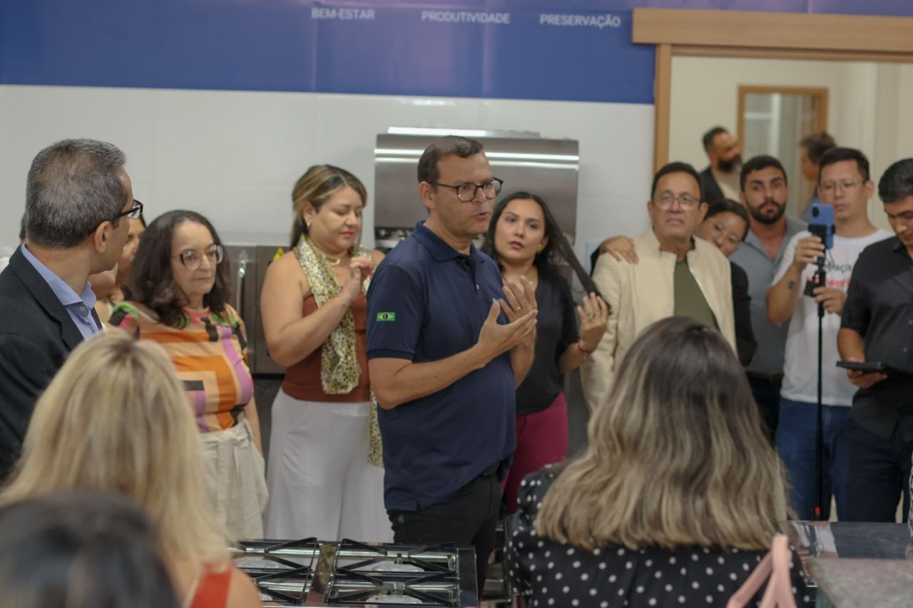 Grupo de pessoas em uma cozinha; um homem de camisa azul fala, enquanto os outros aplaudem.