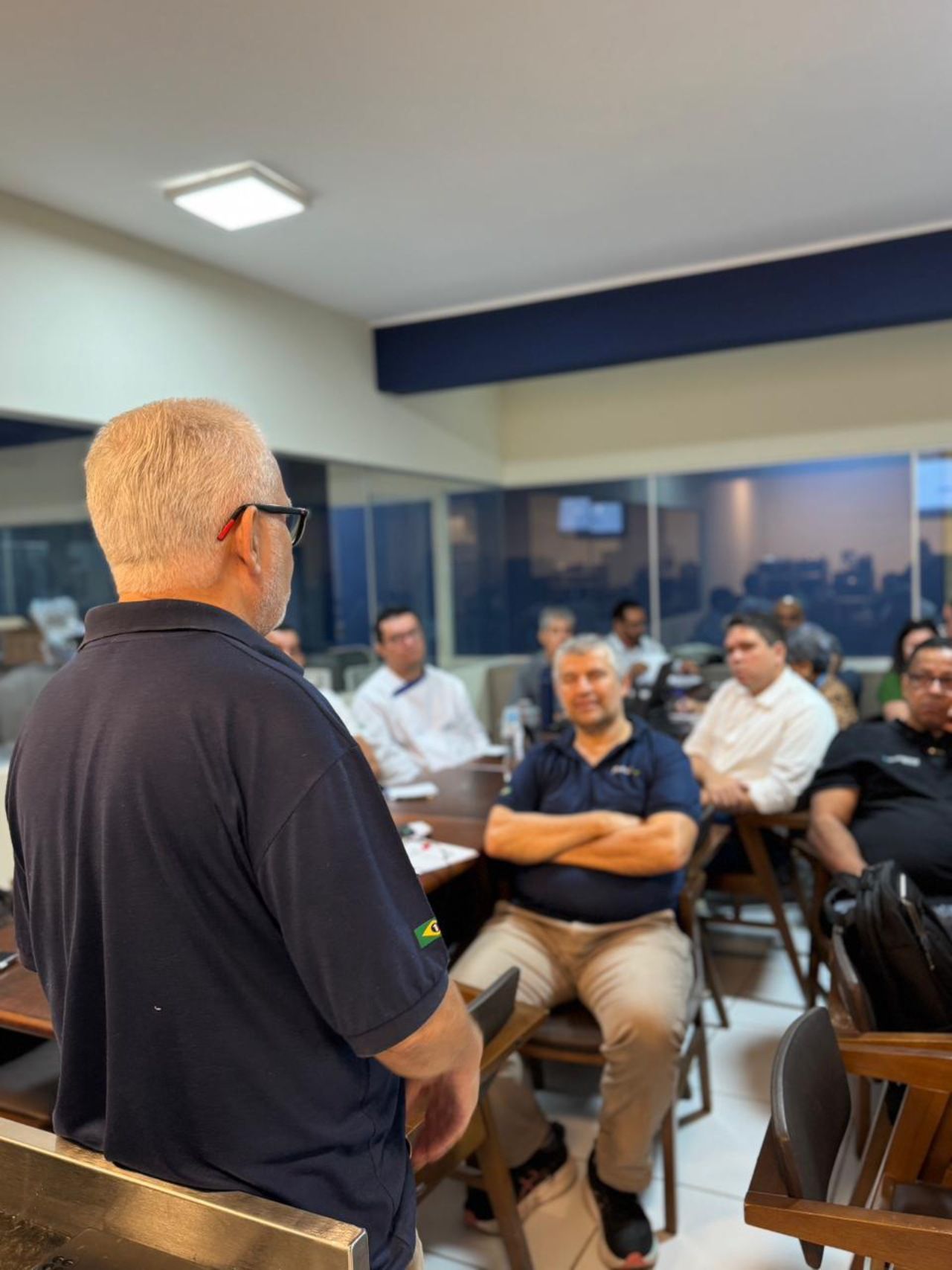 Homem falando para um grupo em uma sala de reuniões. Outros homens sentados, ouvindo. Ambiente interno, bem iluminado.