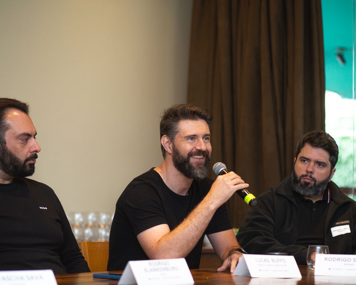 Painel com três homens sentados à mesa. Um deles fala ao microfone, sorrindo. Fundo neutro.