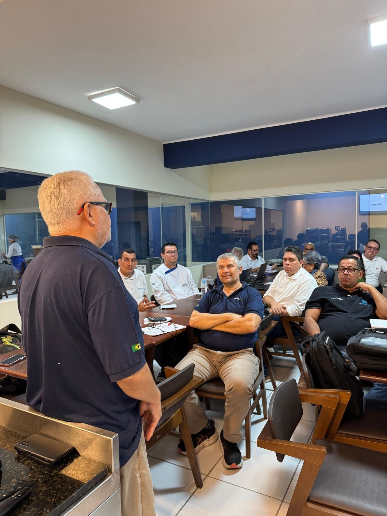 Homem discursando para um grupo em uma sala de reuniões. Os participantes estão sentados às mesas, ouvindo.