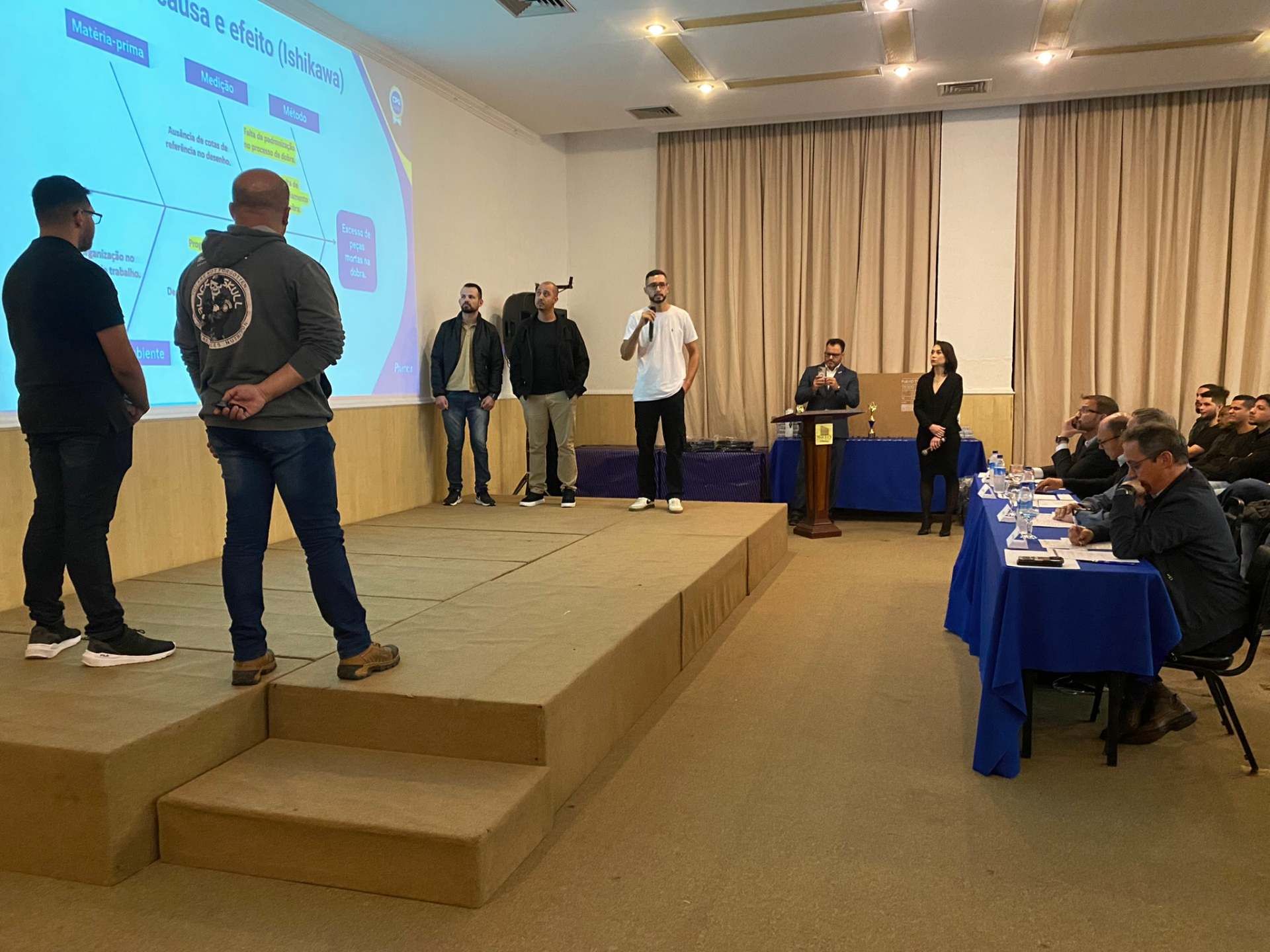 Um grupo de pessoas está em pé em um palco em frente a uma tela grande.