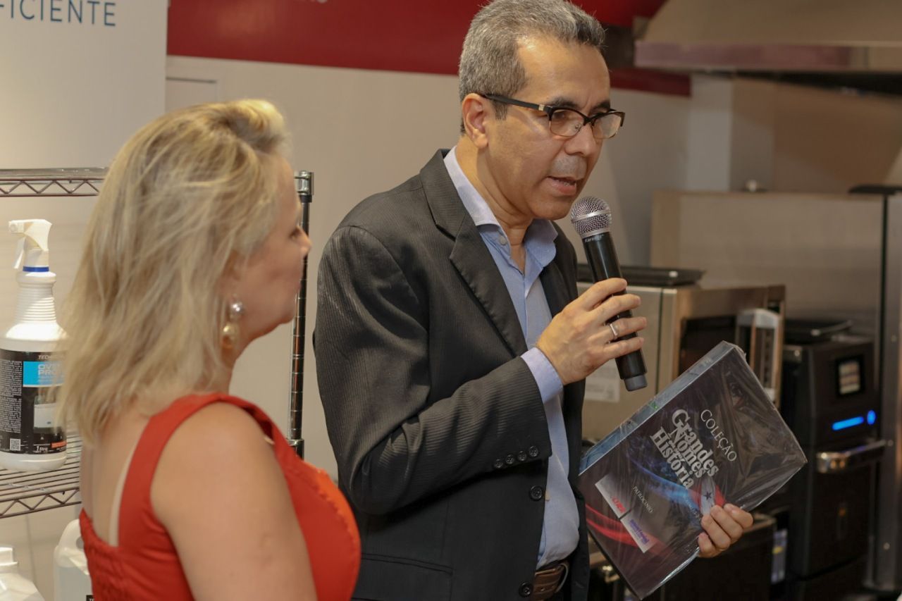 Homem de blazer falando ao microfone, segurando um livro; mulher de vestido vermelho ao seu lado.