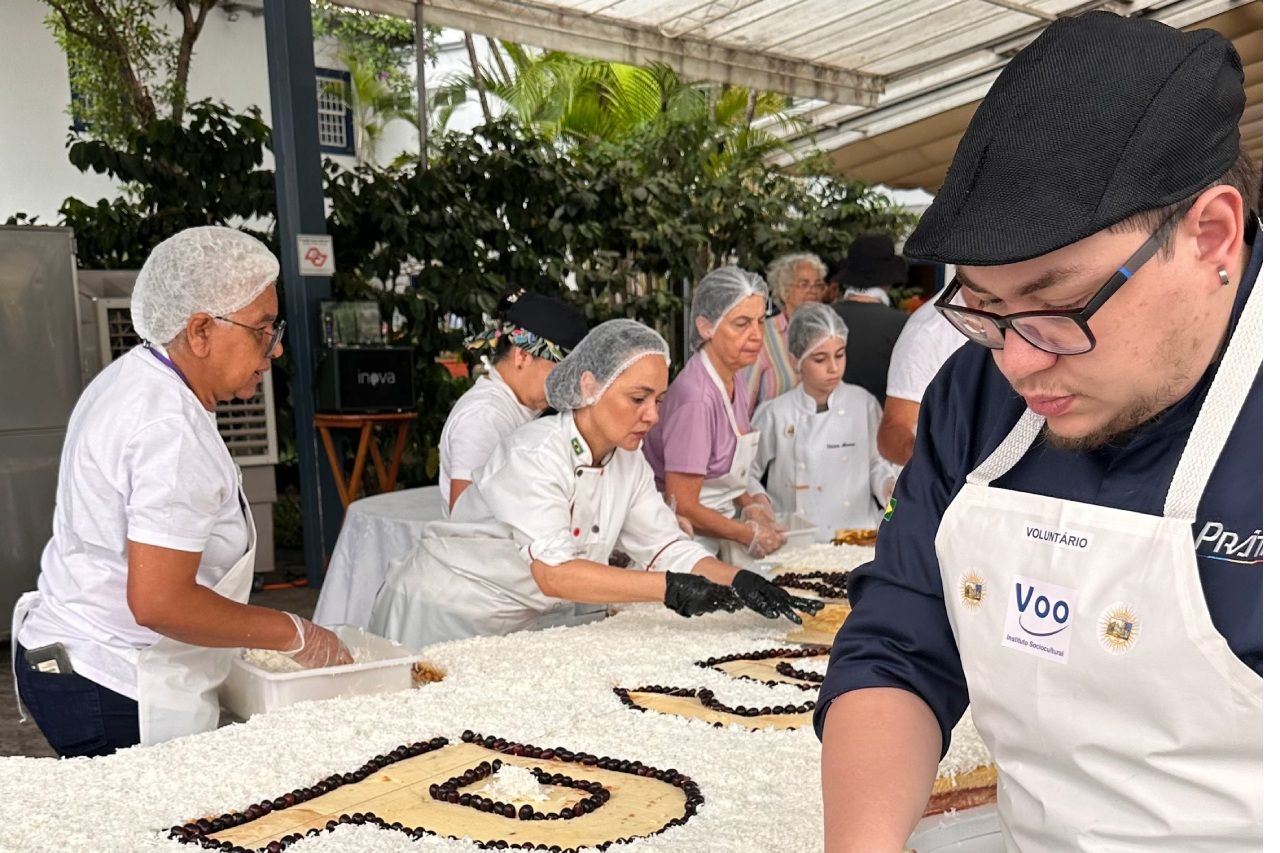 Pessoas decorando um bolo grande ao ar livre, algumas usando aventais e toucas de cabelo.