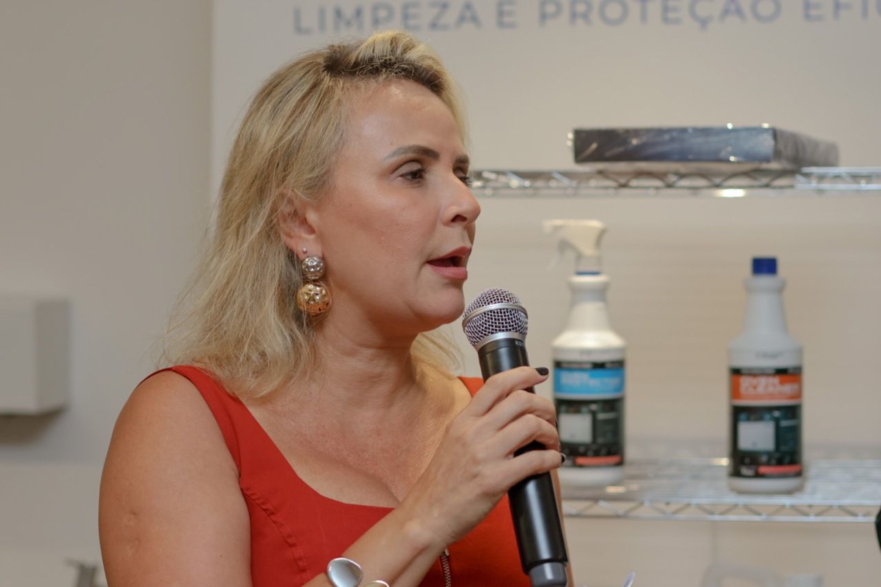 Mulher falando ao microfone, com produtos ao fundo. Cabelo loiro, vestido vermelho, segurando o microfone e falando.