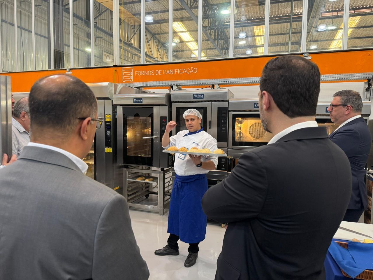 Um chef de avental azul apresentando produtos assados para um grupo em frente aos fornos.