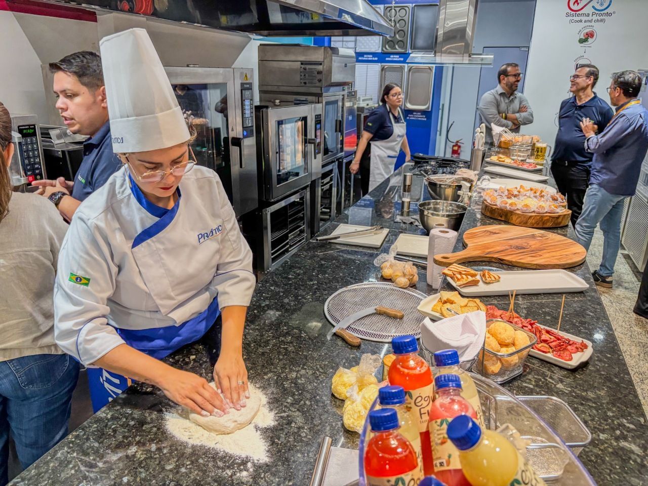 Um chef amassando massa em uma bancada de cozinha; várias pessoas observam.