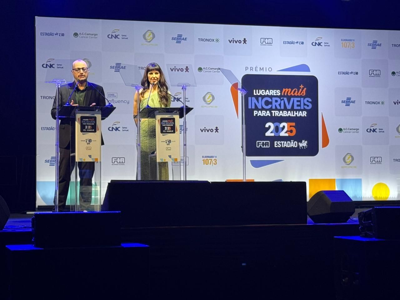 Two people at podiums onstage; event backdrop reads 